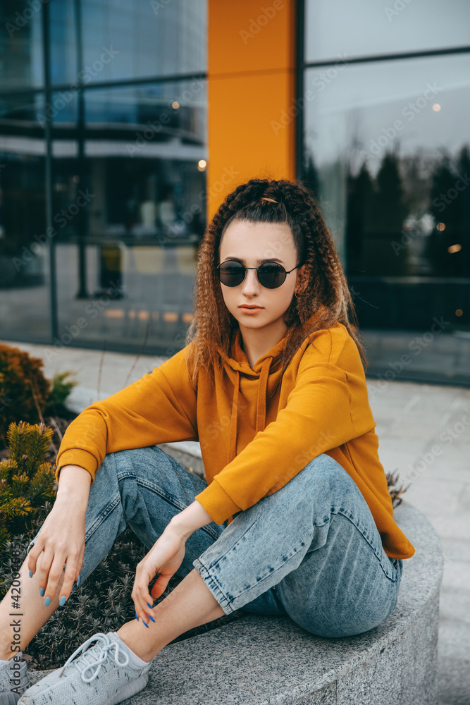 Fototapeta premium Young woman in casual clothes and sunglasses sitting outdoors, on city buildings background, looking at camera.