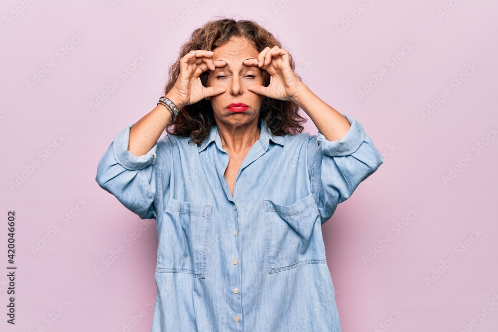 Middle age beautiful woman wearing casual denim shirt standing over pink background Trying to open eyes with fingers, sleepy and tired for morning fatigue Middle age beautiful woman wearing casual denim shirt standing over pink background Trying to open eyes with fingers, sleepy and tired for morning fatigue