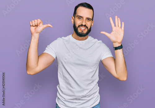 Young man with beard wearing casual white t shirt showing and pointing up with fingers number six while smiling confident and happy.