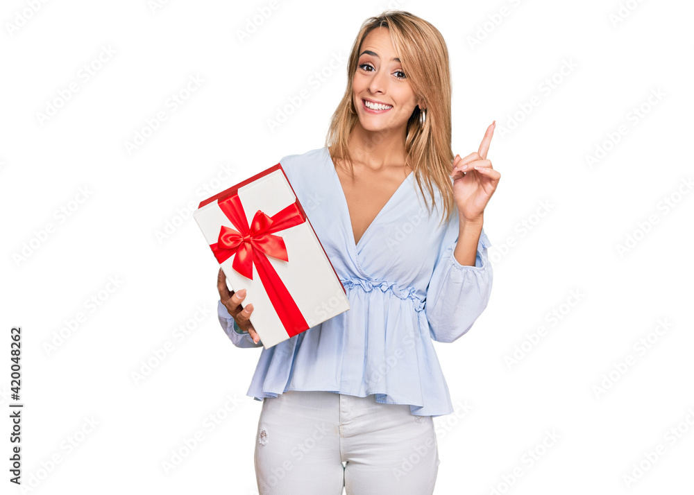 Beautiful blonde woman holding gift surprised with an idea or question pointing finger with happy face, number one Beautiful blonde woman holding gift surprised with an idea or question pointing finger with happy face, number one