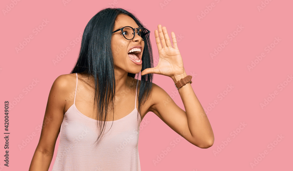 Young african american woman wearing casual clothes and glasses shouting and screaming loud to side with hand on mouth. communication concept. Young african american woman wearing casual clothes and glasses shouting and screaming loud to side with hand on mouth. communication concept.