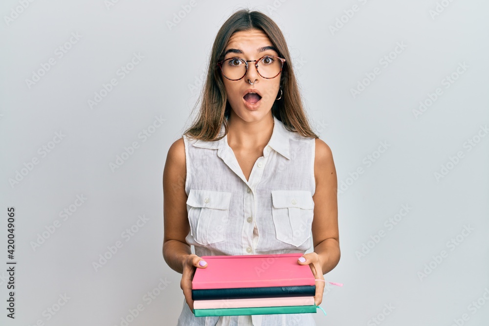 Young hispanic woman holding books afraid and shocked with surprise and amazed expression, fear and excited face. Young hispanic woman holding books afraid and shocked with surprise and amazed expression, fear and excited face.
