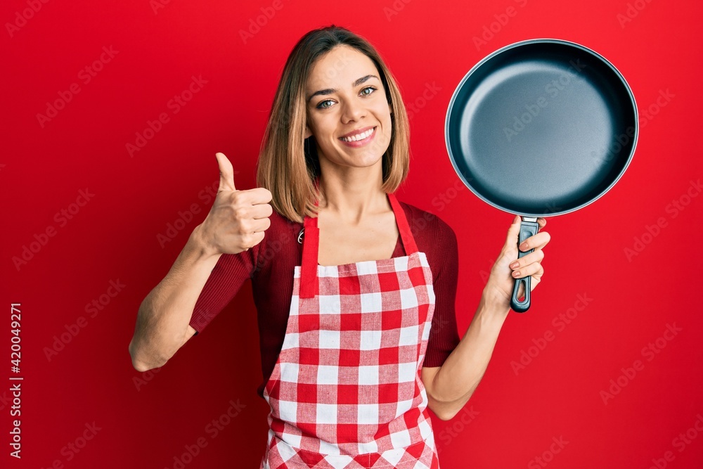 Young caucasian blonde woman wearing cook apron holding pan smiling happy and positive, thumb up doing excellent and approval sign Young caucasian blonde woman wearing cook apron holding pan smiling happy and positive, thumb up doing excellent and approval sign