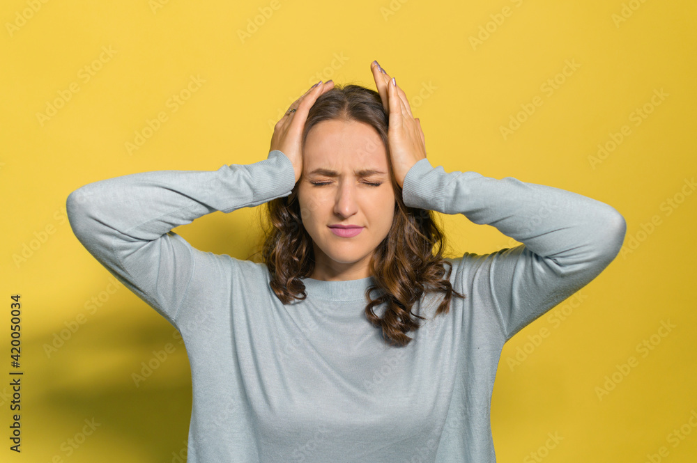 A young woman suffering from headache, holding head with hands, a female closed eyes and feel migraine, feeling stressed and depressed, isolated on yellow A young woman suffering from headache, holding head with hands, a female closed eyes and feel migraine, feeling stressed and depressed, isolated on yellow