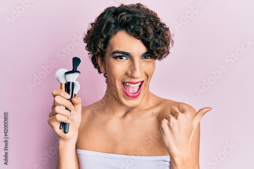 Young man wearing woman make up holding set of brushes pointing thumb up to the side smiling happy with open mouth