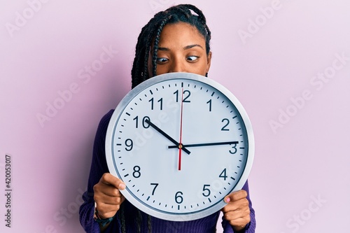 African american woman with braids holding big clock covering face making fish face with mouth and squinting eyes, crazy and comical. photo