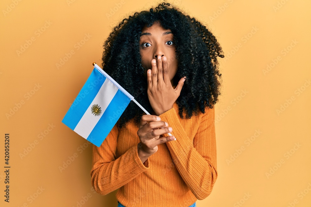African american woman with afro hair holding argentina flag covering mouth with hand, shocked and afraid for mistake. surprised expression African american woman with afro hair holding argentina flag covering mouth with hand, shocked and afraid for mistake. surprised expression