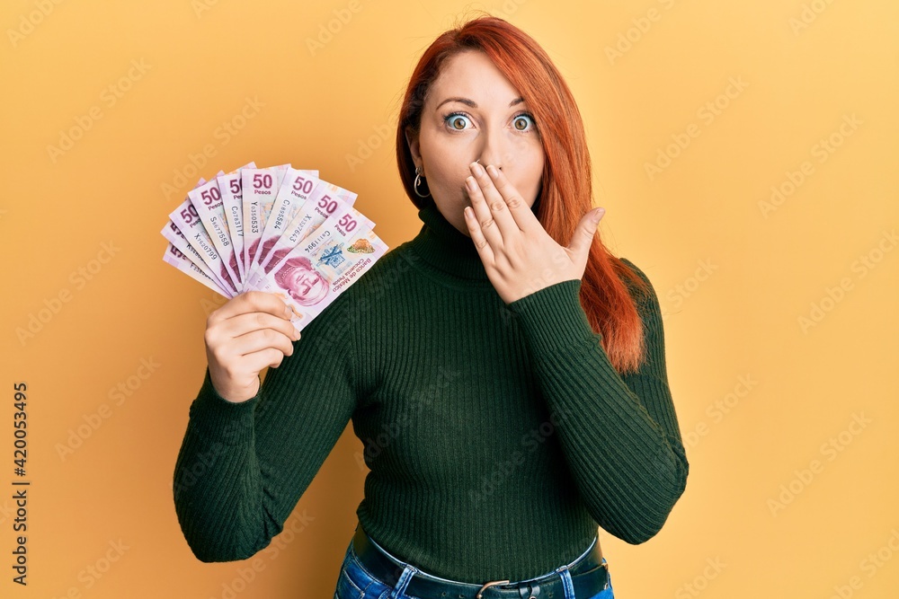 Beautiful redhead woman holding 50 mexican pesos banknotes covering mouth with hand, shocked and afraid for mistake. surprised expression Beautiful redhead woman holding 50 mexican pesos banknotes covering mouth with hand, shocked and afraid for mistake. surprised expression