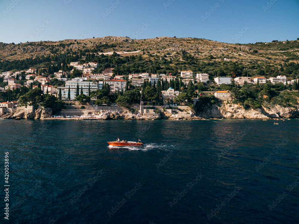Luxurious wooden boat with a motor sails on the background of the coast near the old town of Dubrovnik and Villa Sheherezade. Villas and hotels on the coast of Croatia.