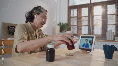 Medium shot of Asian Mature sick patient female talking to laptop screen making video call with male online doctor for discuss health and medicine problems. Senior woman telehealth during lockdown