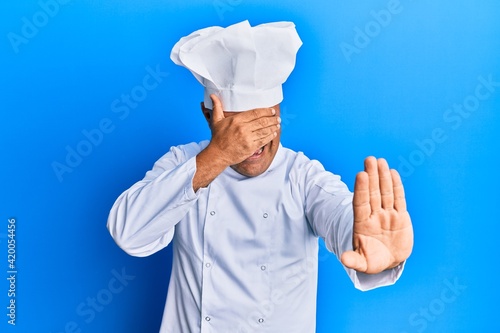 Mature middle east man wearing professional cook uniform and hat covering eyes with hands and doing stop gesture with sad and fear expression. embarrassed and negative concept.