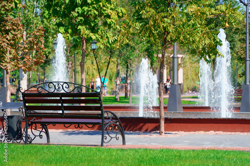 city park on a summer day, green lawns with grass and trees, path, benches and fountain, bright sunlight and shadows