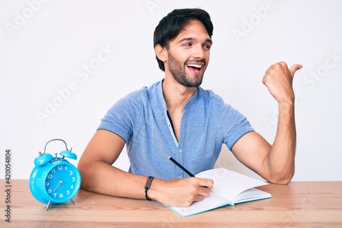 Handsome hispanic man sitting on the table stuying for university pointing thumb up to the side smiling happy with open mouth photo