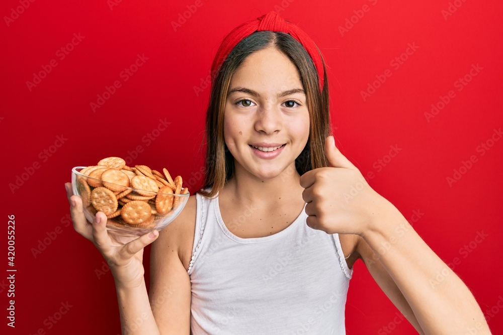 Beautiful brunette little girl holding salty biscuits in a bowl smiling happy and positive, thumb up doing excellent and approval sign Beautiful brunette little girl holding salty biscuits in a bowl smiling happy and positive, thumb up doing excellent and approval sign
