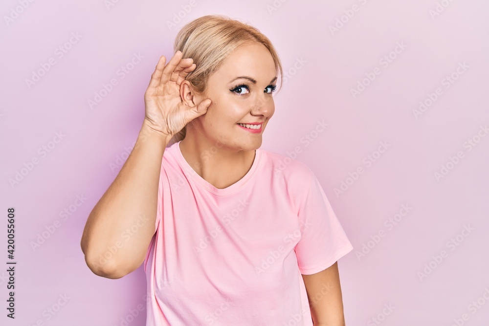 Young blonde woman wearing casual pink t shirt smiling with hand over ear listening an hearing to rumor or gossip. deafness concept. Young blonde woman wearing casual pink t shirt smiling with hand over ear listening an hearing to rumor or gossip. deafness concept.