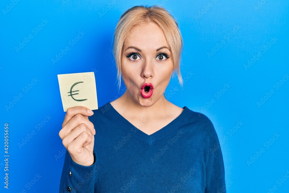 Young blonde woman holding euro symbol reminder scared and amazed with open mouth for surprise, disbelief face Young blonde woman holding euro symbol reminder scared and amazed with open mouth for surprise, disbelief face
