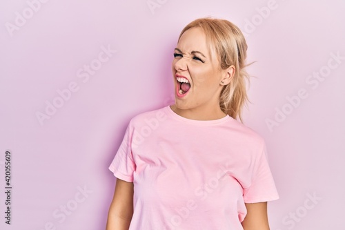 Young blonde woman wearing casual pink t shirt angry and mad screaming frustrated and furious, shouting with anger. rage and aggressive concept.