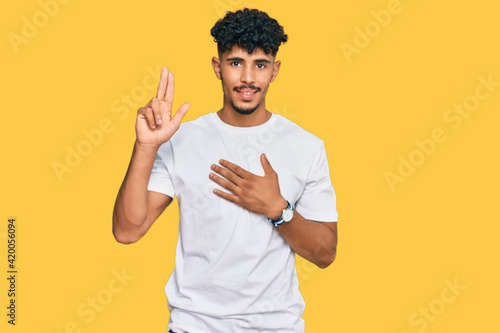 Young arab man wearing casual white t shirt smiling swearing with hand on chest and fingers up, making a loyalty promise oath