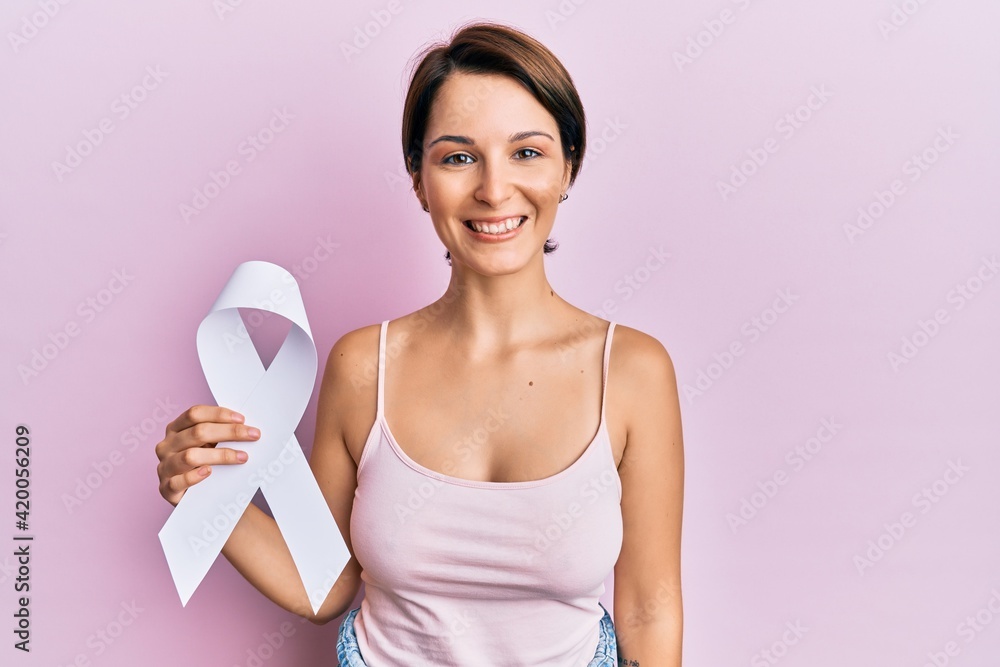 Young brunette woman with short hair holding cancer awareness white ribbon looking positive and happy standing and smiling with a confident smile showing teeth Young brunette woman with short hair holding cancer awareness white ribbon looking positive and happy standing and smiling with a confident smile showing teeth