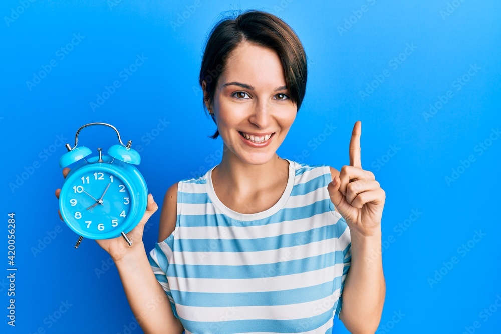 Young brunette woman with short hair holding alarm clock smiling with an idea or question pointing finger with happy face, number one Young brunette woman with short hair holding alarm clock smiling with an idea or question pointing finger with happy face, number one