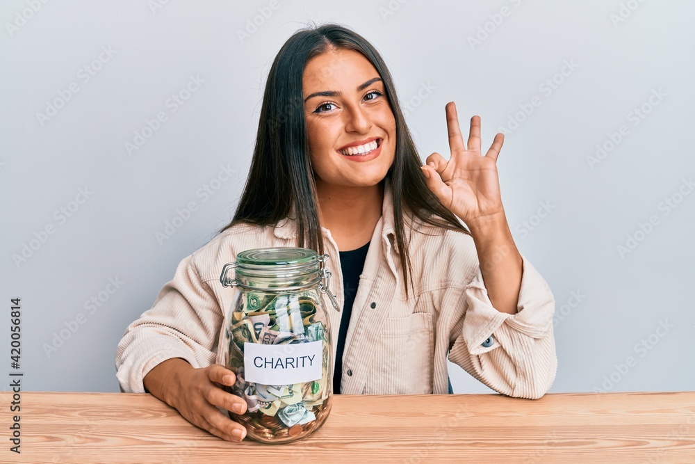 Beautiful hispanic woman holding charity jar with money doing ok sign with fingers, smiling friendly gesturing excellent symbol Beautiful hispanic woman holding charity jar with money doing ok sign with fingers, smiling friendly gesturing excellent symbol