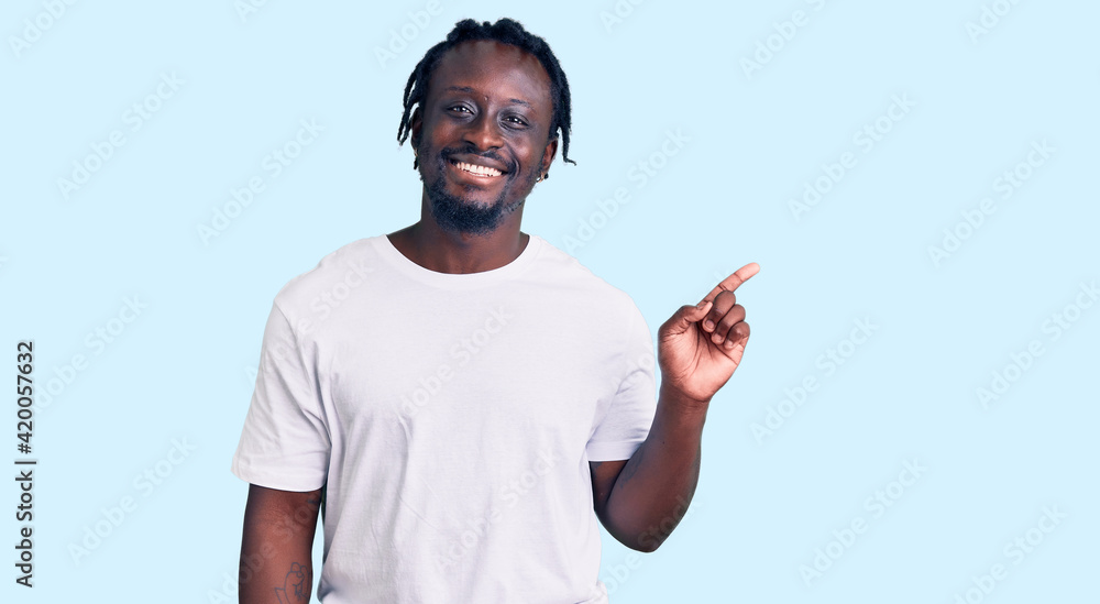 Young african american man with braids wearing casual white tshirt with a big smile on face, pointing with hand finger to the side looking at the camera. Young african american man with braids wearing casual white tshirt with a big smile on face, pointing with hand finger to the side looking at the camera.