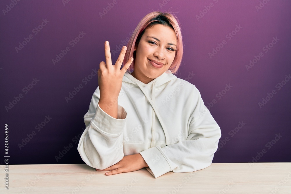 Hispanic woman with pink hair wearing casual sweatshirt sitting on the table showing and pointing up with fingers number three while smiling confident and happy. Hispanic woman with pink hair wearing casual sweatshirt sitting on the table showing and pointing up with fingers number three while smiling confident and happy.