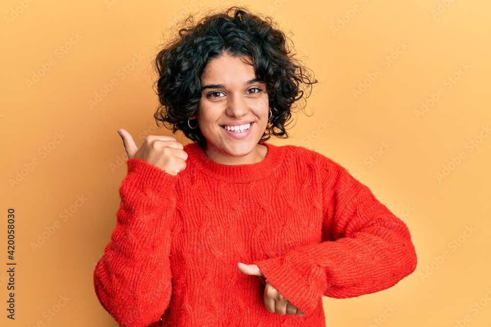Young hispanic woman with curly hair wearing casual winter sweater pointing to the back behind with hand and thumbs up, smiling confident Young hispanic woman with curly hair wearing casual winter sweater pointing to the back behind with hand and thumbs up, smiling confident