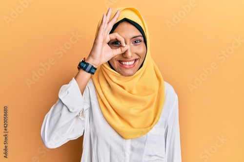 Young brunette arab woman wearing traditional islamic hijab scarf doing ok gesture with hand smiling, eye looking through fingers with happy face. © Krakenimages.com