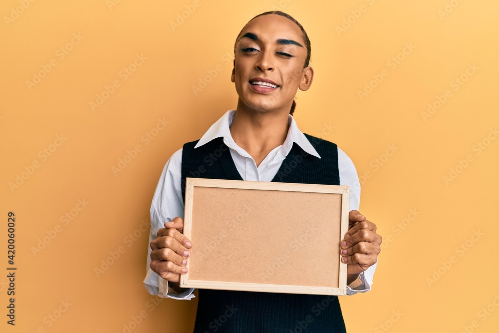 Hispanic man wearing make up and long hair holding empty corkboard winking looking at the camera with sexy expression, cheerful and happy face. Hispanic man wearing make up and long hair holding empty corkboard winking looking at the camera with sexy expression, cheerful and happy face.