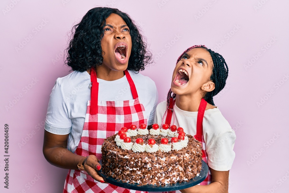 Beautiful african american mother and daughter wearing baker apron holding homemade cake angry and mad screaming frustrated and furious, shouting with anger looking up.