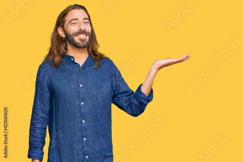 Attractive man with long hair and beard wearing casual clothes smiling cheerful presenting and pointing with palm of hand looking at the camera.