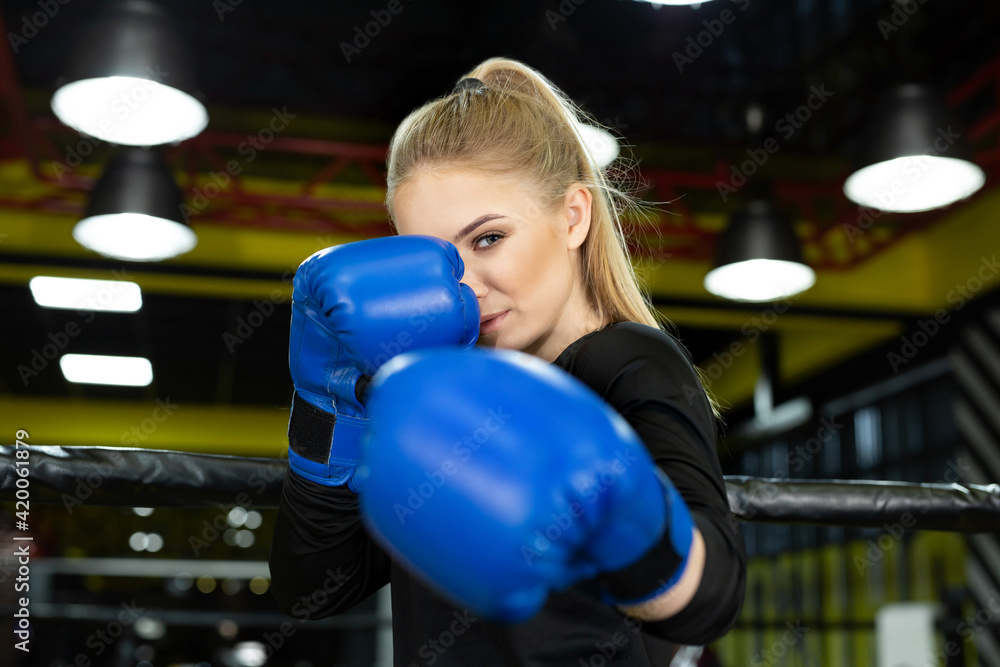 Young beautiful athlete in blue boxing gloves poses in the ring and looks at the camera.