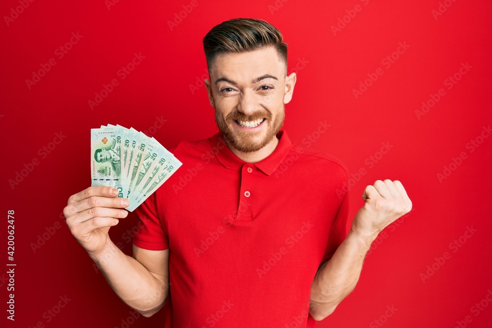 Young redhead man holding 20 thai baht banknotes screaming proud, celebrating victory and success very excited with raised arm Young redhead man holding 20 thai baht banknotes screaming proud, celebrating victory and success very excited with raised arm