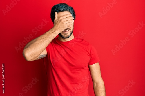 Young hispanic man wearing casual red t shirt smiling and laughing with hand on face covering eyes for surprise. blind concept.