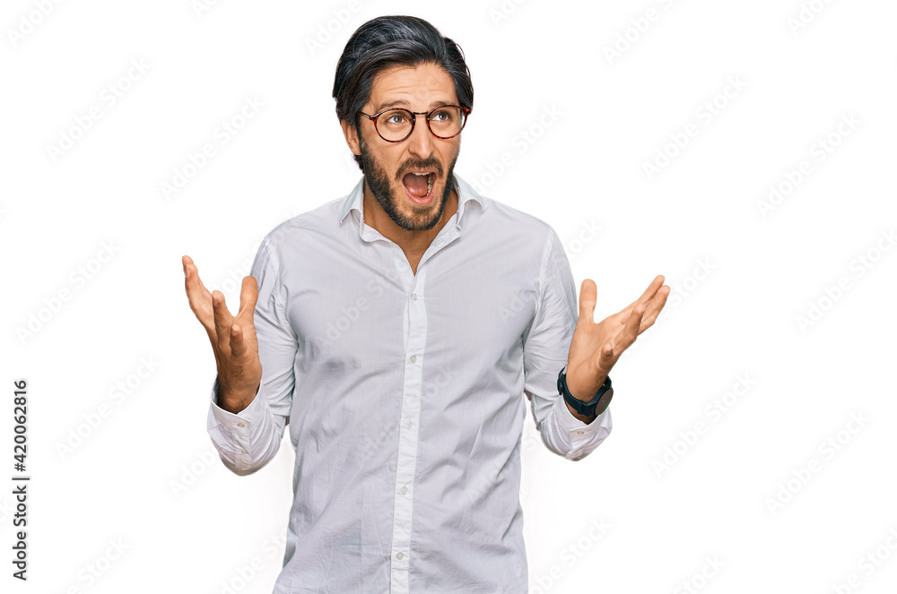 Young hispanic man wearing business shirt and glasses crazy and mad shouting and yelling with aggressive expression and arms raised. frustration concept. Young hispanic man wearing business shirt and glasses crazy and mad shouting and yelling with aggressive expression and arms raised. frustration concept.