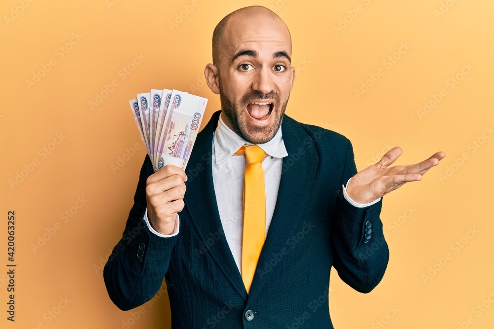 Young hispanic business man holding russian 500 ruble banknotes celebrating achievement with happy smile and winner expression with raised hand Young hispanic business man holding russian 500 ruble banknotes celebrating achievement with happy smile and winner expression with raised hand