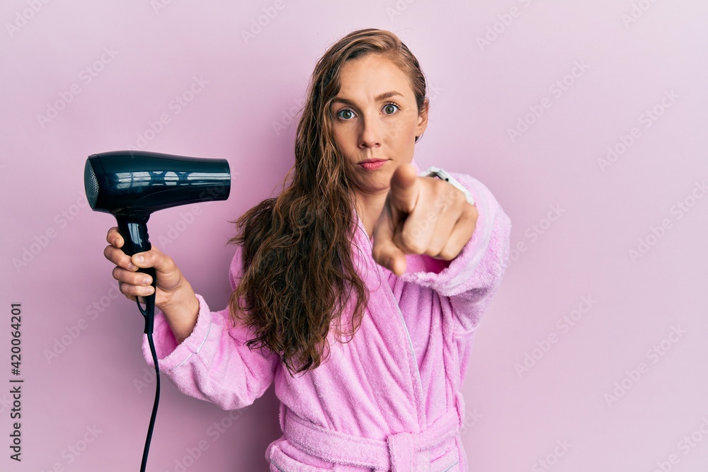 Young blonde woman wearing bathrobe using hair dryer pointing with finger to the camera and to you, confident gesture looking serious Young blonde woman wearing bathrobe using hair dryer pointing with finger to the camera and to you, confident gesture looking serious