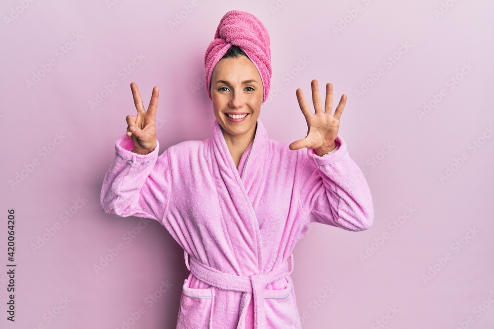 Young blonde woman wearing shower towel cap and bathrobe showing and pointing up with fingers number seven while smiling confident and happy. Young blonde woman wearing shower towel cap and bathrobe showing and pointing up with fingers number seven while smiling confident and happy.