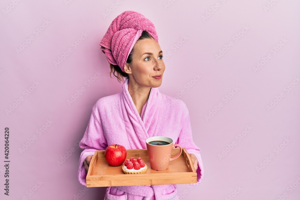Young blonde woman wearing bathrobe holding tray with breakfast smiling looking to the side and staring away thinking. Young blonde woman wearing bathrobe holding tray with breakfast smiling looking to the side and staring away thinking.
