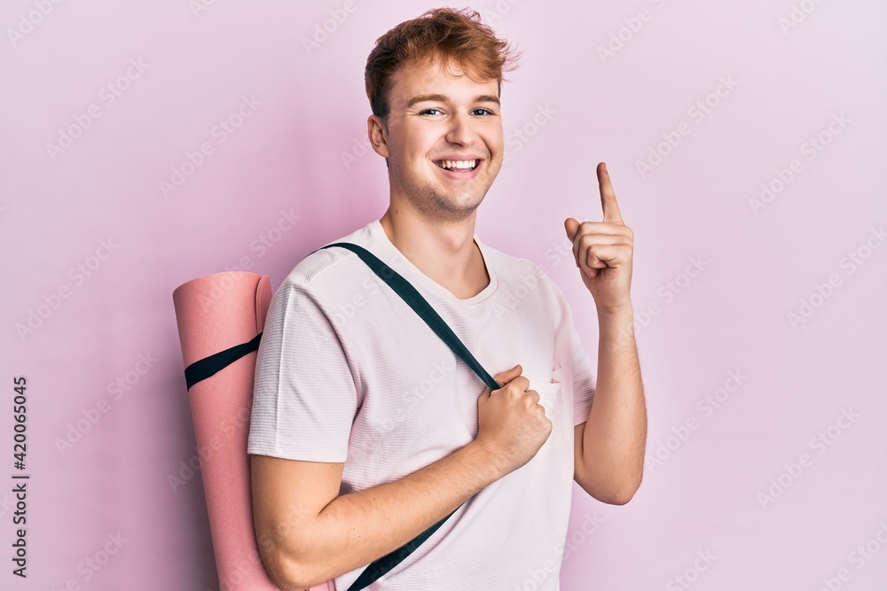 Young caucasian man holding yoga mat smiling with an idea or question pointing finger with happy face, number one Young caucasian man holding yoga mat smiling with an idea or question pointing finger with happy face, number one