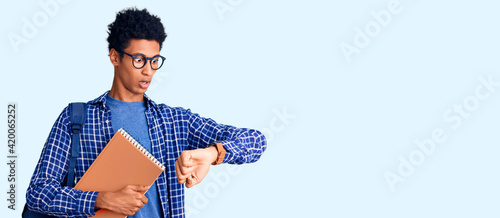 Young african american man wearing student backpack holding book looking at the watch time worried, afraid of getting late photo