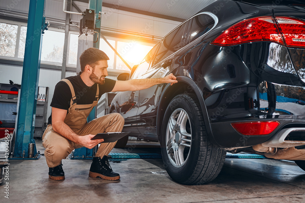 Handsome mechanic in uniform is making notes examining car in auto service. - obrazy, fototapety, plakaty 