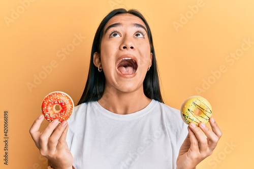 Young asian woman holding tasty colorful doughnuts angry and mad screaming frustrated and furious  shouting with anger looking up.