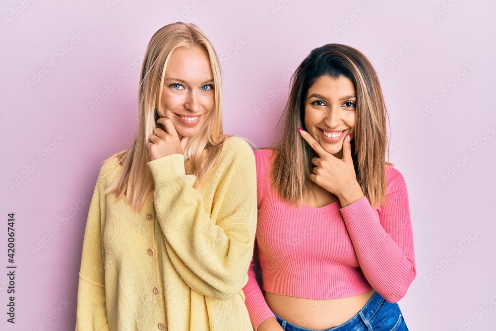 Two friends standing together over pink background looking confident at the camera smiling with crossed arms and hand raised on chin. thinking positive. Two friends standing together over pink background looking confident at the camera smiling with crossed arms and hand raised on chin. thinking positive.