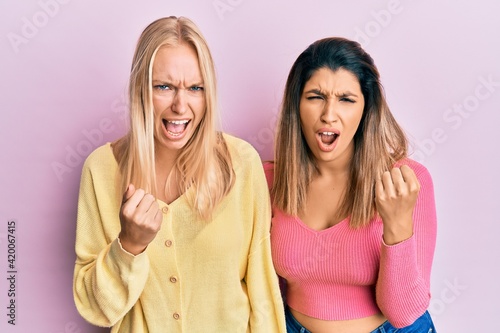Two friends standing together over pink background angry and mad raising fist frustrated and furious while shouting with anger. rage and aggressive concept.
