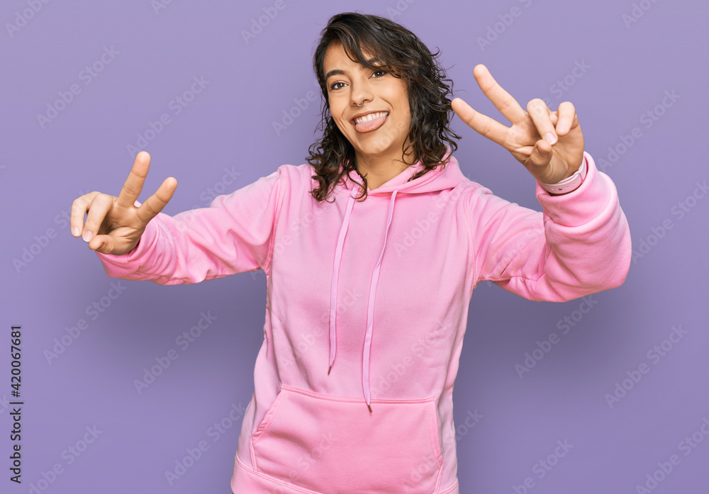 Young hispanic woman wearing casual sweatshirt smiling with tongue out showing fingers of both hands doing victory sign. number two. Young hispanic woman wearing casual sweatshirt smiling with tongue out showing fingers of both hands doing victory sign. number two.