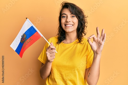 Young hispanic woman holding russia flag doing ok sign with fingers  smiling friendly gesturing excellent symbol