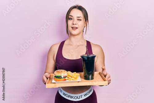 Young hispanic girl wearing sportswear eating a tasty classic burger with fries and soda winking looking at the camera with sexy expression, cheerful and happy face.