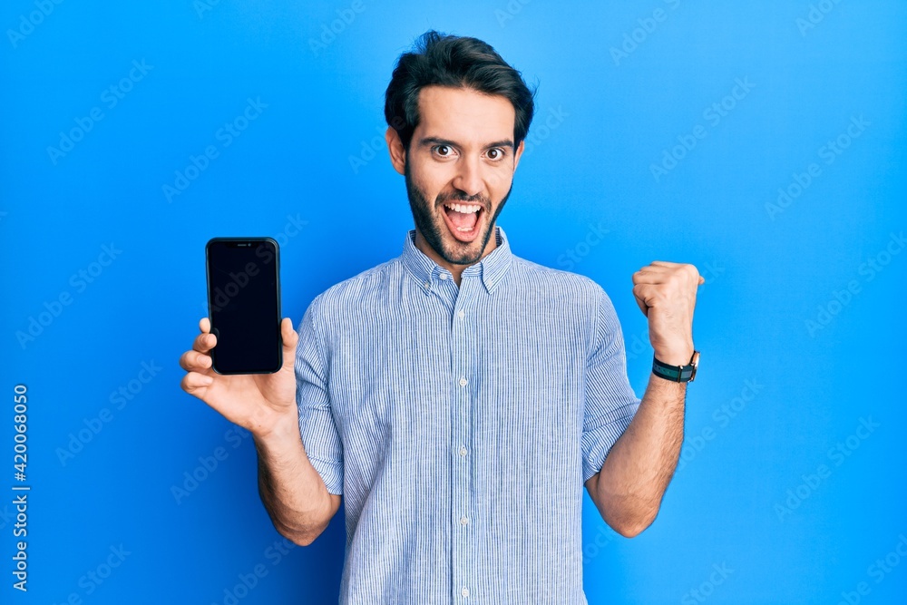Young hispanic man holding smartphone showing screen pointing thumb up to the side smiling happy with open mouth Young hispanic man holding smartphone showing screen pointing thumb up to the side smiling happy with open mouth